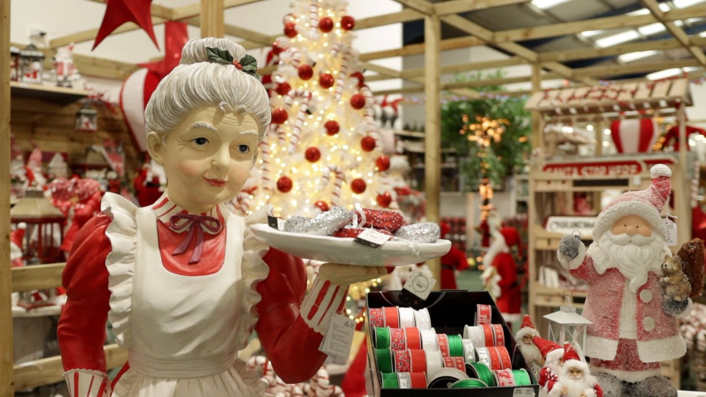 Mrs Claus holding tray with Santa and white Christmas Tree with red decorations in the background